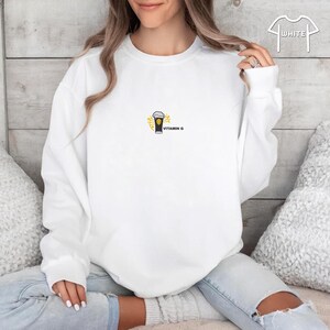 May include: White sweatshirt with a crew neck. The front features a small graphic of a beer glass with foam, surrounded by a yellow sunburst design. The text "VITAMIN G" is below the graphic. The sweatshirt is a casual style.