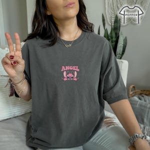 May include: Dark gray t-shirt with a pink graphic of a cartoon character and the word "ANGEL" above it. The character has large eyes and ears. The shirt is a comfortable, oversized fit.