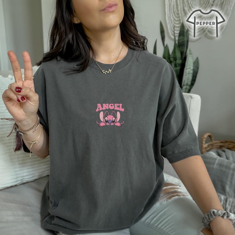 May include: Dark gray t-shirt with a pink graphic of a cartoon character and the word "ANGEL" above it. The character has large eyes and ears. The shirt is a comfortable, oversized fit.