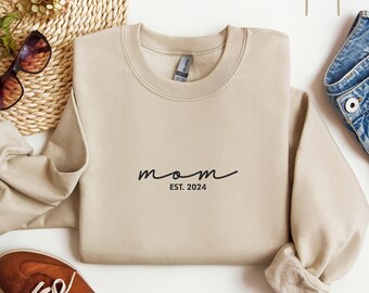 May include: A beige sweatshirt with the text "mom est. 2024" in black.