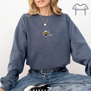 May include: A denim blue sweatshirt with a small embroidered design of a beer glass and the words "VITAMIN G". The sweatshirt has long sleeves and a crew neck. The model is wearing blue jeans and black sneakers.