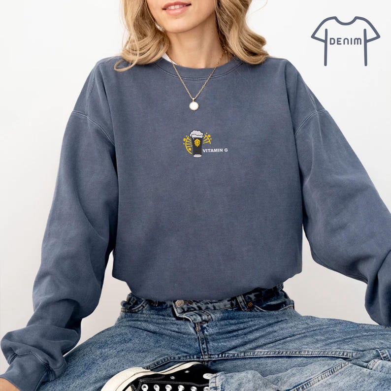 May include: A denim blue sweatshirt with a small embroidered design of a beer glass and the words "VITAMIN G". The sweatshirt has long sleeves and a crew neck. The model is wearing blue jeans and black sneakers.