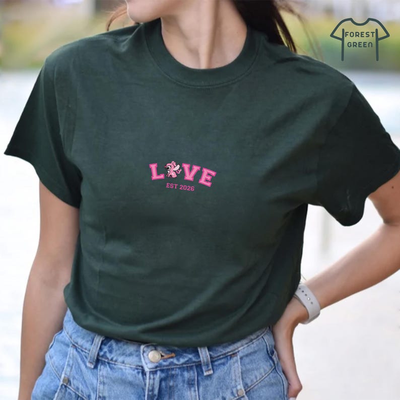 Puede incluir: Camiseta verde oscuro con la palabra "LOVE" bordada en rosa, con un dise&ntilde;o floral y el texto "EST 2026". La camiseta tiene mangas cortas y cuello redondo. El logo "FOREST GREEN" est&aacute; en la esquina superior derecha.