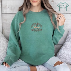 May include: A light green sweatshirt with a brown graphic of a book with a feather and the text "Flourish & Blots Booksellers".