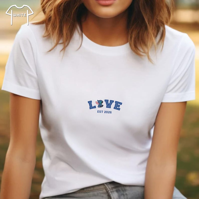 Puede incluir: Camiseta blanca con un dise&ntilde;o bordado en azul que dice "LOVE" con un personaje de dibujos animados y el texto "EST 2026". La camiseta tiene mangas cortas y cuello redondo.