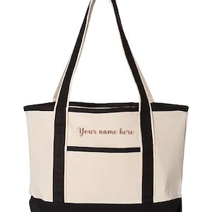 May include: Cream-colored tote bag with black accents and handles. The bag features a front pocket and the text "Your name here" in a script font. The bottom of the bag is black.
