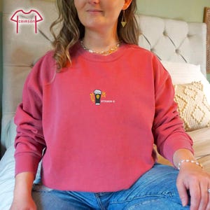 May include: A crimson-colored sweatshirt with a crew neck. The front features a small graphic of a beer mug with the text "VITAMIN C" in white. The sweatshirt is made of a soft material and is suitable for casual wear.
