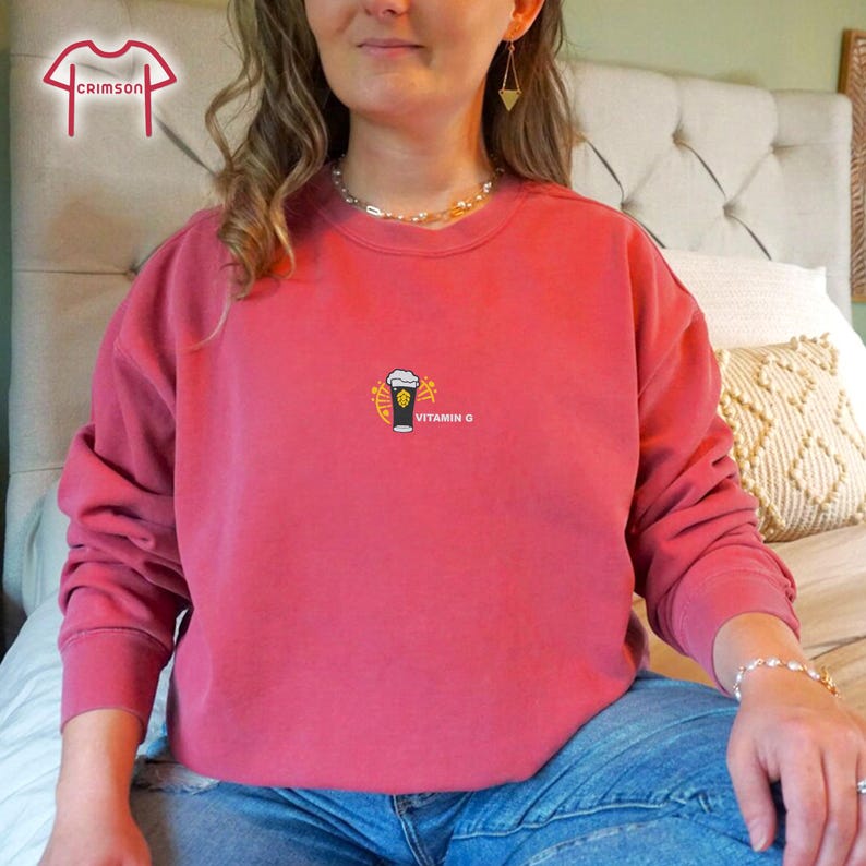 May include: A crimson-colored sweatshirt with a crew neck. The front features a small graphic of a beer mug with the text "VITAMIN C" in white. The sweatshirt is made of a soft material and is suitable for casual wear.