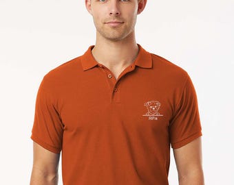 May include: A burnt orange polo shirt with a white embroidered dog graphic and the name "Alfie" underneath. The shirt has a classic collar and short sleeves. The model is wearing a black belt.