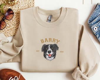 May include: Beige sweatshirt with the name "BARRY" in gold lettering above a black and white dog illustration. The text "EST 2024" is below the dog. The sweatshirt has a crew neck and long sleeves.