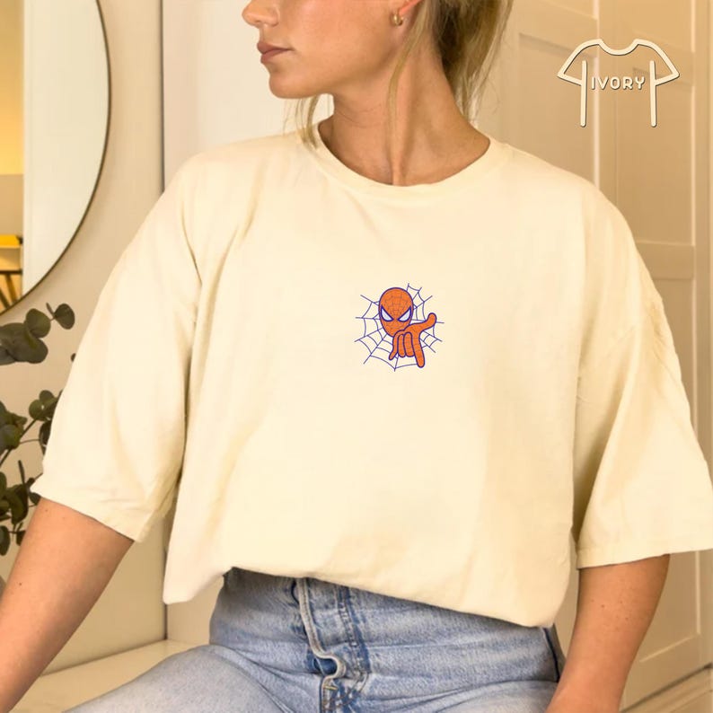May include: A cream colored t-shirt with a cartoon image of Spiderman in a web. The image is in orange and blue.