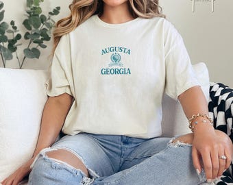 May include: A white t-shirt with a vintage-style graphic that says "Augusta, Georgia" with a crest featuring a pine tree and the words "Founded 1735".