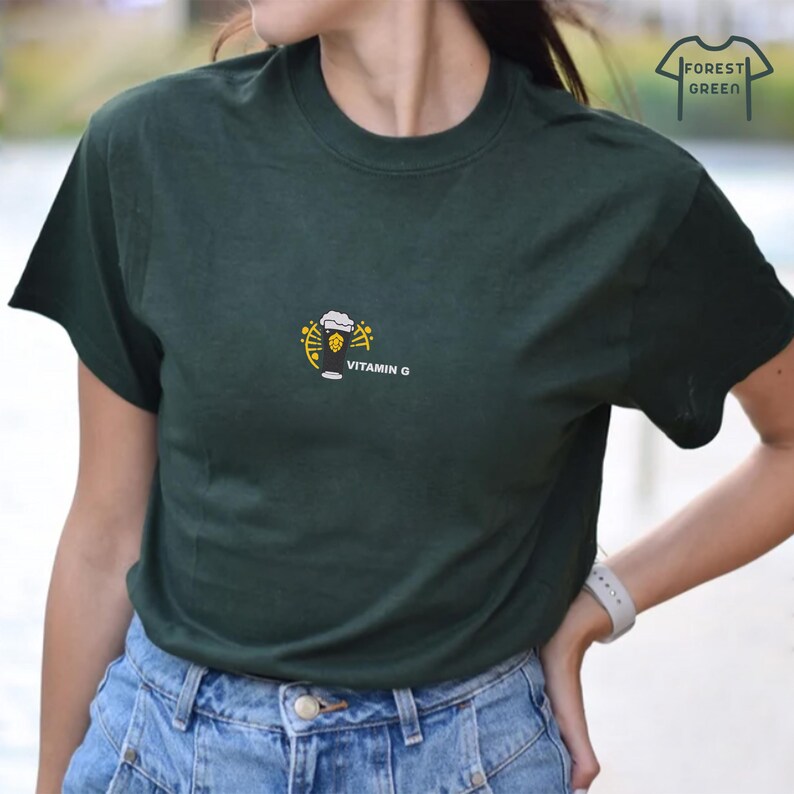 Puede incluir: Camiseta verde oscuro con un peque&ntilde;o dise&ntilde;o bordado de una jarra de cerveza y el texto "VITAMIN G" debajo. La camiseta tambi&eacute;n tiene un peque&ntilde;o logotipo que dice "FOREST GREEN". La camiseta est&aacute; hecha de un material suave.