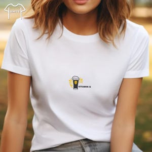Puede incluir: Camiseta blanca con un peque&ntilde;o gr&aacute;fico de un vaso de cerveza con espuma y el texto "VITAMIN G". El vaso de cerveza es negro y amarillo, con un dise&ntilde;o de explosi&oacute;n amarilla alrededor.