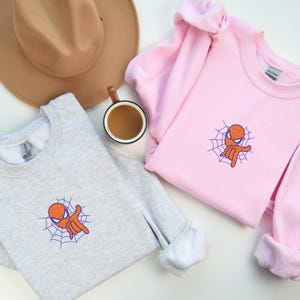 May include: Two sweatshirts with a cartoon image of Spiderman giving a peace sign. The sweatshirt on the left is gray and the sweatshirt on the right is pink.