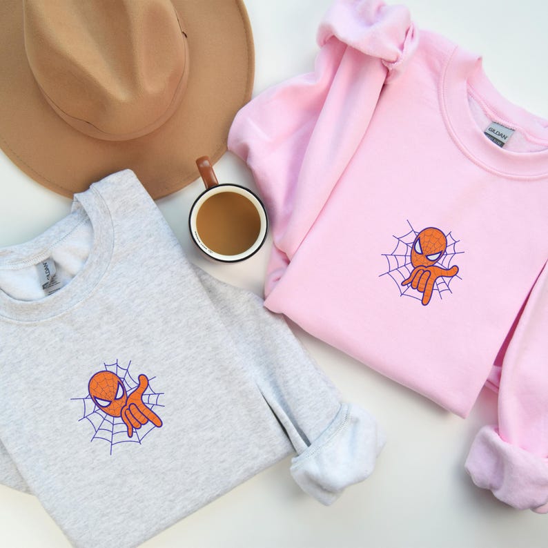May include: Two sweatshirts with a cartoon image of Spiderman giving a peace sign. The sweatshirt on the left is gray and the sweatshirt on the right is pink.