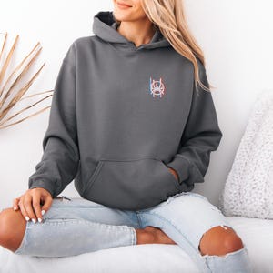 K&ouml;nnte beinhalten: Ein anthrazitfarbener Kapuzenpullover mit einer Vordertasche und einer Kapuze. Der Kapuzenpullover hat ein kleines, farbenfrohes Emblem auf der oberen linken Brust. Die Person tr&auml;gt hellblaue Jeans mit zerrissenen Knien.