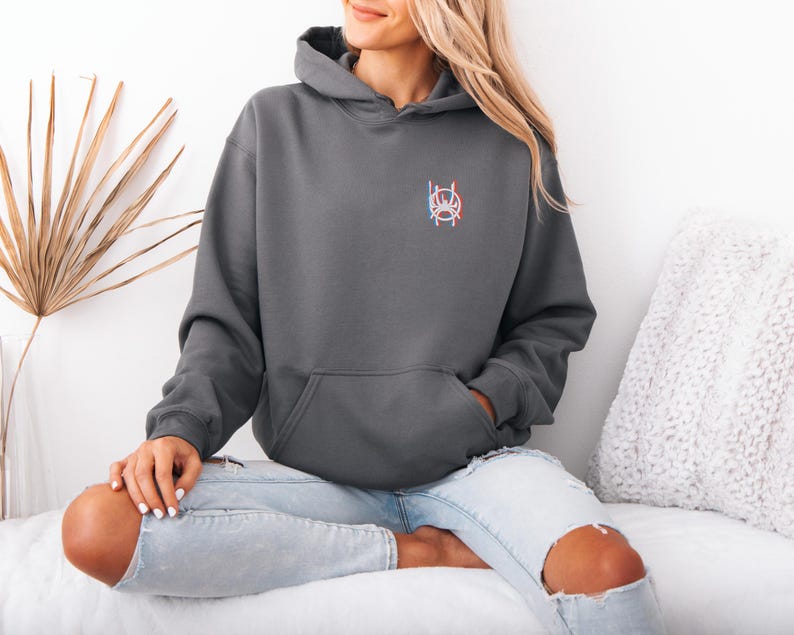 K&ouml;nnte beinhalten: Ein anthrazitfarbener Kapuzenpullover mit einer Vordertasche und einer Kapuze. Der Kapuzenpullover hat ein kleines, farbenfrohes Emblem auf der oberen linken Brust. Die Person tr&auml;gt hellblaue Jeans mit zerrissenen Knien.