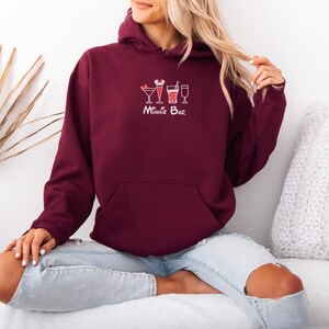 May include: A maroon hoodie with a front pocket and a drawstring hood. The hoodie features a white graphic of three cocktails and the text "Minnie Bar" in a script font. The person is wearing blue jeans.
