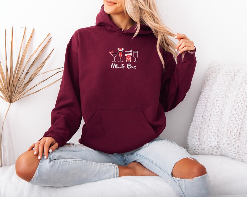 May include: A maroon hoodie with a front pocket and a drawstring hood. The hoodie features a white graphic of three cocktails and the text "Minnie Bar" in a script font. The person is wearing blue jeans.
