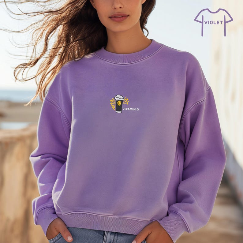 May include: A violet crewneck sweatshirt with a small embroidered graphic of a beer glass with the text "VITAMIN G". The sweatshirt has a relaxed fit and long sleeves. The word "VIOLET" is printed above the sweatshirt.