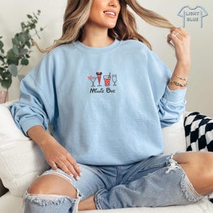 Puede incluir: Una sudadera de color azul claro con un gr&aacute;fico de Minnie Mouse y una variedad de bebidas. El texto "Minnie Bar" est&aacute; impreso debajo del gr&aacute;fico.