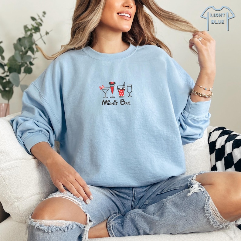 Puede incluir: Una sudadera de color azul claro con un gr&aacute;fico de Minnie Mouse y una variedad de bebidas. El texto "Minnie Bar" est&aacute; impreso debajo del gr&aacute;fico.