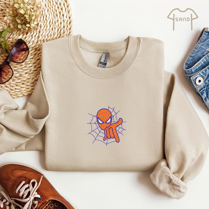 May include: A beige sweatshirt with a cartoon image of Spiderman in a web. The image is in orange, blue, and white.