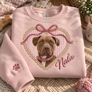 Peut inclure: Un sweat-shirt à col rond rose clair avec un portrait brodé d'un chien marron portant un collier rose. Le chien est encadré par un cœur avec un nœud rose et le nom Nala brodé en écriture cursive. Une petite empreinte de patte est sur la manche.
