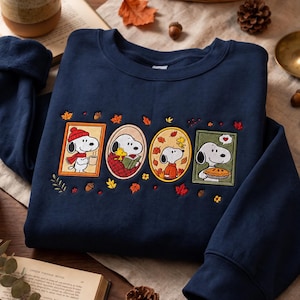 Peut inclure: Un sweat-shirt bleu marine à col rond orné de broderies sur le thème de l'automne représentant Snoopy et Woodstock. Le motif comprend quatre illustrations encadrées des personnages avec des feuilles, des glands et une tarte.