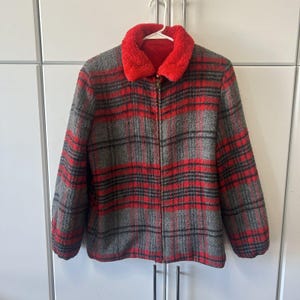 May include: A zip-up jacket featuring a red and gray plaid pattern with a textured, fuzzy finish. The garment includes a bright red faux fur collar, long sleeves, and a central metal zipper closure that extends from the hem to the neckline.