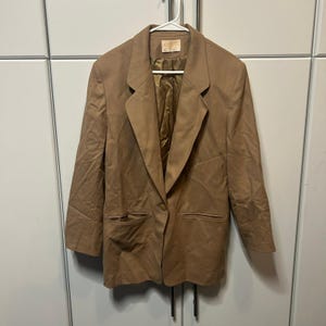 May include: A tan blazer featuring a single-button closure, notched lapels, and two front welt pockets. The jacket includes long sleeves, a structured collar, and a smooth, matching tan interior lining. A small rectangular brand label is visible.