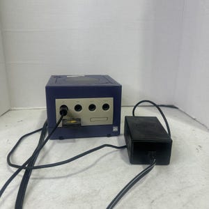Può includere: Una console per videogiochi viola e grigio chiaro con quattro porte per controller sul pannello frontale, una scheda di memoria inserita nel primo slot e un alimentatore nero collegato tramite un lungo cavo nero. La console presenta un logo ATI.