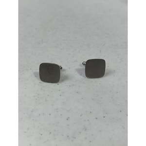 May include: A pair of square cufflinks with a brushed metal finish. The cufflinks have a rounded post on the back for securing to a shirt cuff. The color is a muted silver-gray.