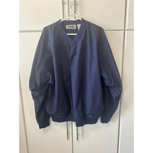 Op de afbeelding: Een marineblauwe windjack met een V-hals en elastische manchetten en zoom. De jas hangt aan een witte hanger. Het woord "TOUR" is op de kraag geborduurd.