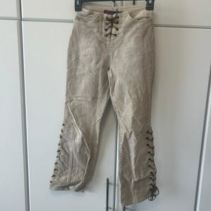 May include: Beige corduroy pants featuring a high-waisted design with a central lace-up closure. The pants include dark brown laces that thread through metal eyelets at the waist and along the outer seams of both pant legs from the knee down.