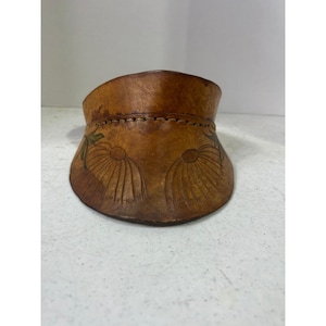 May include: A brown leather pouch with a curved top and a stitched seam. The pouch features embossed floral designs and green accents. The leather has a natural, aged appearance.
