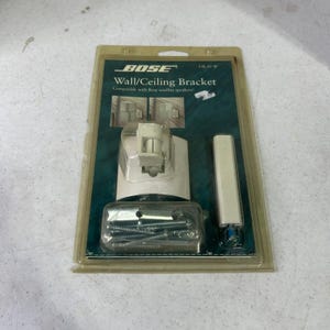 May include: A Bose UB-20 W wall and ceiling bracket in original plastic packaging. The kit includes a white mounting base, a rectangular extension arm, and a small clear plastic bag containing metal screws and hardware for speaker installation.