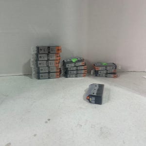 May include: A collection of rectangular printer ink cartridges in various stacks. The cartridges are primarily grey and black with orange accents and printed labels including the numbers 220 and 221. One individual cartridge sits separately in the foreground.