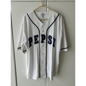 May include: White baseball jersey with navy blue trim and the word "PEPSI" in navy blue block letters across the chest. The jersey has a v-neck, button-up front, and short sleeves. The jersey is hanging on a wooden hanger.