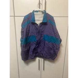 May include: A long-sleeve windbreaker jacket featuring a color-blocked design in deep purple and teal. The jacket has a full-length front zipper, a high collar, and a white interior lining. The fabric appears lightweight with a crinkled texture.