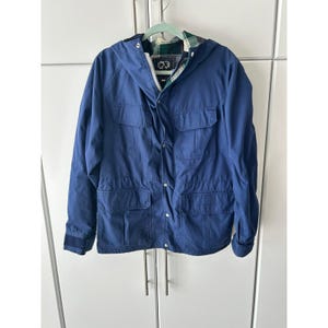 May include: A navy blue hooded jacket with multiple front pockets and a drawstring waist. The jacket features a plaid lining in the hood and a zipper closure. The jacket is hanging on a light green hanger against a white background.
