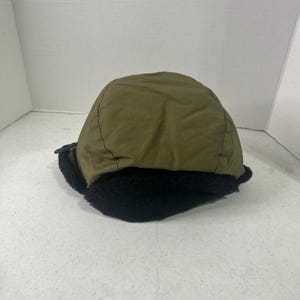 May include: An olive green winter hat featuring a smooth, solid-colored fabric crown and a thick, black faux fur lining along the bottom edge. The hat has a rounded shape with visible stitched seams and a dark green cord detail on the side.