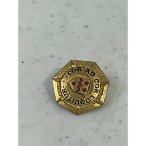 May include: A gold-colored, octagonal pin with the words "COR AD COR LOQUITUR" around the edge. The center features a shield design with red accents. The pin is likely a vintage or antique item.