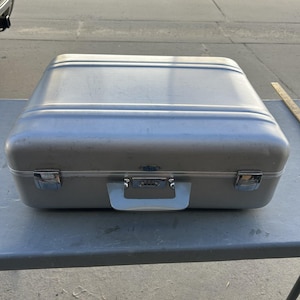 May include: A silver-colored rectangular hard-shell carrying case with a metallic finish, featuring a central handle, two side latches, and a small blue logo emblem on the front. A wooden ruler is visible on the right side of the case.
