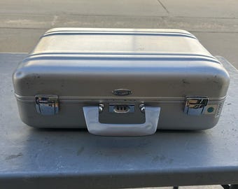 May include: A rectangular silver hard-shell carrying case with a smooth metallic finish. It features a central handle, two chrome-plated metal latches, and a combination lock mechanism. A small label on the side displays the text VX1000.
