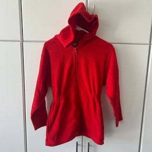 May include: A bright red zip-up hoodie made of a textured fleece material. The garment features long sleeves, a hood, and a cinched waistline with an elasticized band. The front includes a full-length zipper closure that extends from the hem to the neck.