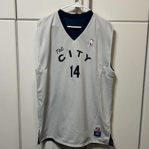 May include: A sleeveless basketball jersey in light gray with a dark navy blue V-neck collar. The front features the text The CITY in navy blue lettering above the number 14. An NBA logo is on the upper right chest and a small rectangular patch is at the hem.