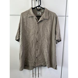 Puede incluir: Una camisa de manga corta con botones en color verde oliva apagado. La camisa tiene cuello y un sutil estampado floral en relieve. Cuelga de una percha negra sobre un fondo blanco.
