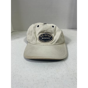 Op de afbeelding: Een beige baseballpet met een gebogen klep. De pet toont de tekst "US OPEN 2000 PEBBLE BEACH" in een cirkelvormig embleem op de voorkant. De pet heeft een licht versleten uiterlijk.